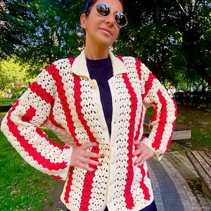 May include: A cream-colored, open-weave crochet cardigan with bold red vertical stripes. The cardigan has a collared neckline and button closures. The garment is worn over a black top and paired with black leggings. The background features green trees and a park setting.