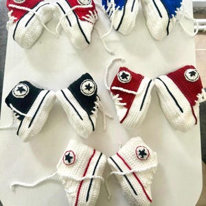 Może przedstawiać: Sześć par szydełkowych bucików dla niemowląt w różnych kolorach, w tym czerwonym, niebieskim, czarnym i białym. Każdy bucik ma białą gwiazdkę na palcach i białą podeszwę.