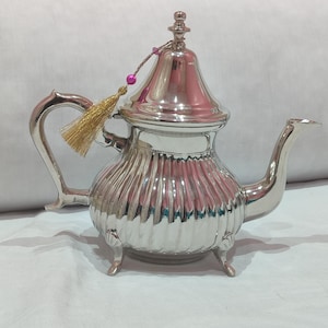 以下が含まれることがあります： リブ加工のボディ、曲がったハンドル、長い注ぎ口が付いた銀色のティーポット。蓋には装飾的なつまみとピンクのビーズが付いたタッセルが付いています。ティーポットは3つの小さな足の上にあります。
