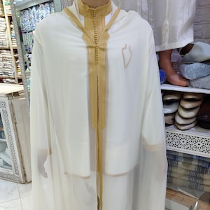 May include: Off-white traditional garment with gold trim. The outfit includes a long, flowing robe and matching pants. The robe features gold embroidery around the neckline and down the front. The outfit is displayed on a mannequin.