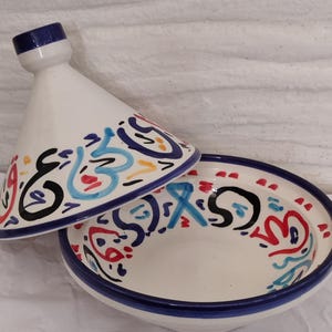 May include: A white ceramic tagine with a conical lid and a round base. The tagine features colourful abstract designs in blue, red, and black. The rim and the top of the lid are trimmed with blue.