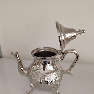 Puede incluir: Una tetera plateada con tapa abierta. La tetera tiene un pico curvo, un asa y está sostenida por tres patas pequeñas. El cuerpo de la tetera está decorado con un patrón floral en relieve.