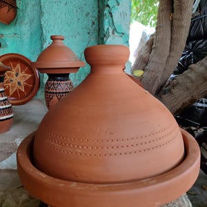 May include: A terracotta tagine pot with a conical lid and a wide, shallow base. The pot has a series of small, evenly spaced holes around the upper portion. Other terracotta pottery is visible in the background.