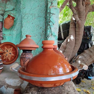 May include: A collection of terracotta tagine pots and serving dishes. The tagine pot in the foreground has a rounded lid and a silver band around the base. Other pots and dishes are visible in the background, some with decorative patterns.