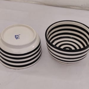 Peut inclure: Deux petits bols en céramique blanche avec des rayures horizontales noires. L'un des bols est à l'endroit, l'autre est à l'envers, révélant un tampon bleu. Les bols ont un design simple et moderne.