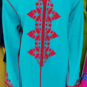 May include: A turquoise hooded robe with red embroidery featuring a floral design. The robe has a zipper closure and a long, flowing silhouette.