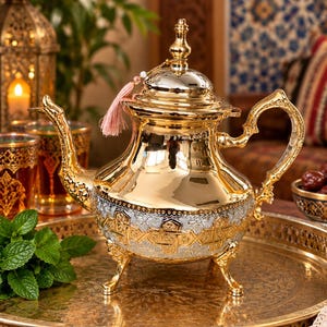 May include: A polished gold and silver teapot featuring an ornate embossed geometric pattern on the body, a curved handle, a long spout, and three decorative feet. A small pink tassel is attached to the lid, which has a pointed finial top.
