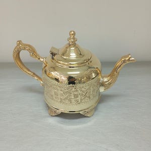 Könnte beinhalten: Eine goldene Teekanne mit einem verzierten Design und einem Scharnierdeckel. Die Teekanne hat einen gebogenen Ausguss und einen Griff mit einem dekorativen Knauf.