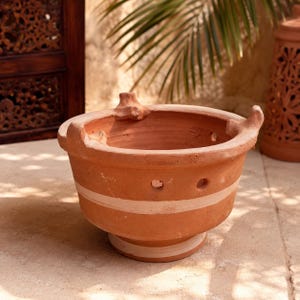 May include: A terracotta clay pot with a wide circular opening, featuring three small protruding handles on the rim. The exterior has a row of small circular ventilation holes and two horizontal white painted stripes around the body and base.