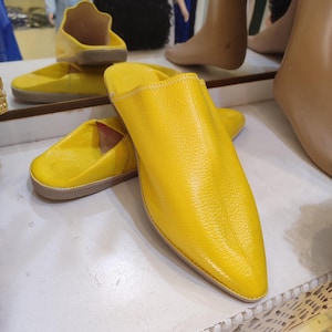 Puede incluir: Zapatillas de cuero amarillo brillante con punta y tacón bajo. Las zapatillas se exhiben sobre una superficie blanca, con otro par visible en el fondo. Las zapatillas tienen un diseño sencillo y clásico.