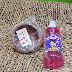 Könnte beinhalten: Eine transparente Plastikflasche mit rosa Flüssigkeit, neben einer kleinen, runden Holzschale, die in transparente Folie eingewickelt ist. Die Flasche hat ein blaues und violettes Etikett mit arabischem Text und einen weißen Sprühkopf.