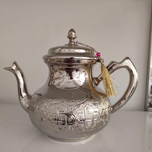 Puede incluir: Tetera plateada con intrincados diseños en relieve. Tiene un pico curvo, un asa y una tapa con un pequeño pomo. Una borla dorada con una cuenta rosa cuelga de la tapa. Ideal para servir té.