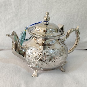 Puede incluir: Una tetera plateada con intrincados patrones en relieve y un asa pequeña. La tetera tiene una tapa con un adorno decorativo y una borla azul. Se apoya sobre tres patas pequeñas.