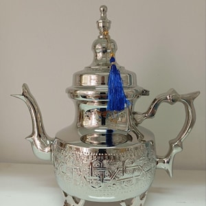 Puede incluir: Una tetera marroquí de plata con intrincados diseños en relieve y un borla azul. La tetera tiene un cuerpo redondo, un pico curvo y una tapa con un pomo.