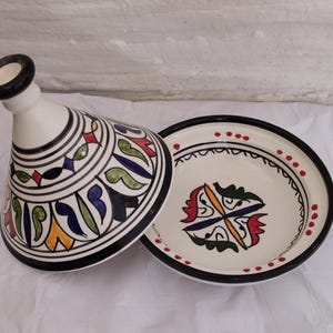 May include: A white ceramic tagine with a conical lid and a round base. The lid and base are decorated with black, blue, green, red, and yellow geometric patterns. The rim of the base has red dots.