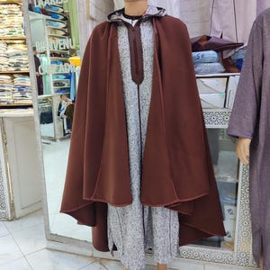 May include: A mannequin displays a brown hooded cape over a long, patterned robe. The robe has a white and gray pattern with a central vertical design. A maroon fez-style hat completes the outfit. The background shows a shop interior.