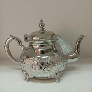 Può includere: Una teiera d'argento con un designo ornato e un fiocco. La teiera ha un corpo rotondo con un coperchio e un beccuccio. Si appoggia su tre gambe.
