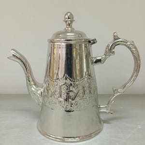 Könnte beinhalten: Eine silberne Teekanne mit einem verzierten Design und einem Scharnierdeckel. Die Teekanne hat einen gebogenen Ausguss und einen Griff mit einem dekorativen Rollendesign.
