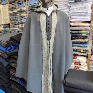 May include: A gray hooded cloak with a textured center panel and a black zipper, displayed on a mannequin. The cloak is paired with a yellow and white patterned head covering. The background shows stacks of folded fabric.