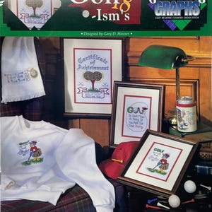 May include: A collection of golf-themed items, including a white sweatshirt with an embroidered golfer, framed cross-stitch art, a red cap, and a mug. The image also features golf balls, tees, and a green desk lamp. The text "Golf ism's" is visible.