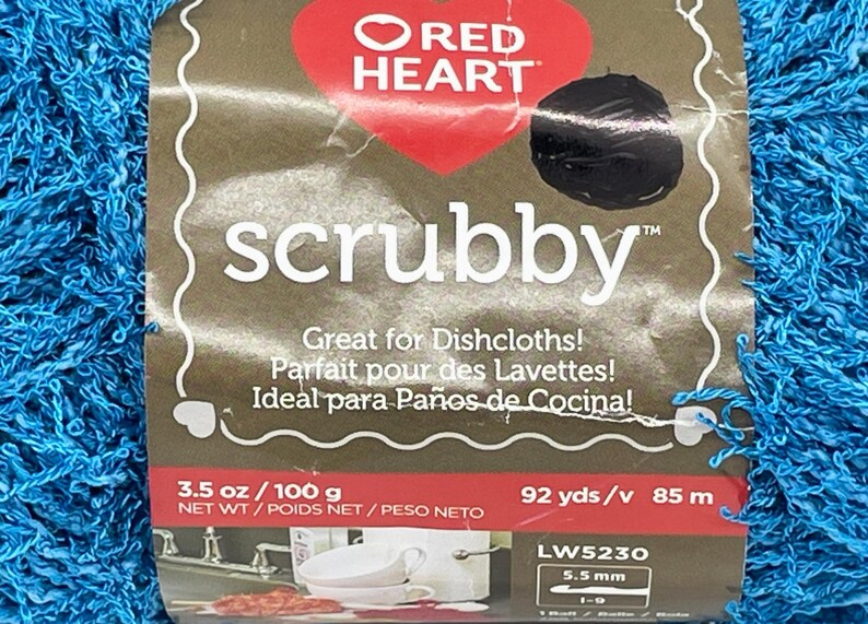 Pode incluir: Um close-up de uma bola de fio azul scrubby. A etiqueta diz "Red Heart scrubby" e inclui texto em ingl&ecirc;s, franc&ecirc;s e espanhol. O fio tem 100 g e 85 m.