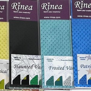May include: A collection of Rinea brand craft paper in various colors, including white, yellow, black, teal, blue, and purple. Each paper pack has a unique texture and is labeled with a descriptive name such as "Magical" and "Patriotic". The Rinea logo and website are visible.