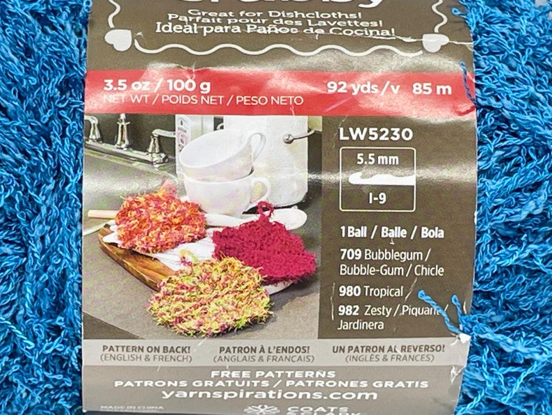 Pode incluir: Um pacote de fio azul com um r&oacute;tulo mostrando panos de prato de croch&ecirc; coloridos. O r&oacute;tulo inclui o texto "Great for Dishcloths!" e medidas de 100 g e 85 m.