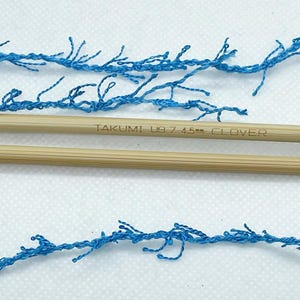 Pode incluir: Close-up de fio azul com textura de franja, disposto com duas agulhas de tric&ocirc; de madeira. As agulhas s&atilde;o castanhas claras e t&ecirc;m o texto "TAKUMI US 7 4.5mm CLOVER" impresso. O fio e as agulhas est&atilde;o sobre uma superf&iacute;cie branca.