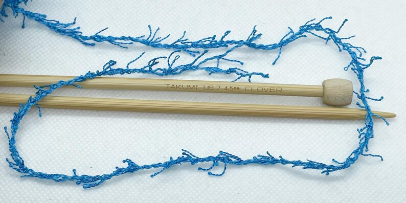 Pode incluir: Close-up de fio azul com textura de franja, disposto com duas agulhas de tric&ocirc; de madeira. As agulhas s&atilde;o castanhas claras e t&ecirc;m o texto "TAKUMI US 7 4.5mm CLOVER" impresso. O fio e as agulhas est&atilde;o sobre uma superf&iacute;cie branca.