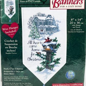 May include: A counted cross-stitch banner kit with a cozy home theme. The design features a snow-covered house, birds, and the text "All hearts come home for Christmas." Includes a wire hanger and tassel. Measures 8" x 14".