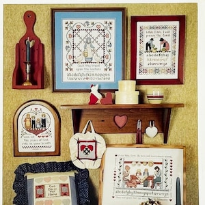 May include: A display of framed cross-stitch samplers and decorative items. The pieces include a framed sampler with the text "Cast thy bread upon the waters", a shelf with a heart cutout, and a small tote bag with a heart design. The theme is folk art.