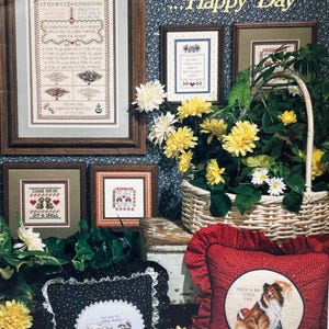 May include: A display of framed cross-stitch samplers and pillows, featuring floral and animal motifs. The text "Sunny Morning...Happy Day" is visible. The collection includes a basket of yellow flowers and a red pillow.