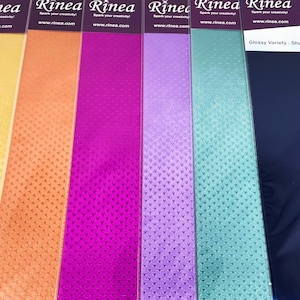 May include: A collection of colorful, glossy paper sheets in shades of yellow, orange, pink, purple, teal, and navy blue. Each sheet has a textured pattern and the brand name "Rinea" is visible at the top.
