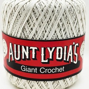 May include: A cylindrical spool of off-white crochet thread wrapped in a tight, crisscross pattern. A red paper label encircles the center, featuring the black text AUNT LYDIA'S and Giant Crochet, with a small circular illustration on each side.
