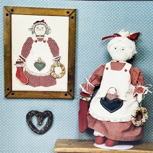 May include: A fabric doll wearing a red and white checkered dress with a white apron, holding a red stocking and a small wreath. Beside it is a framed cross-stitch embroidery of the same doll, and a small dark brown woven heart ornament.