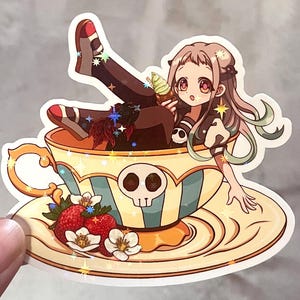 Puede incluir: Una pegatina con un personaje de estilo anime en una taza de té. La taza de té tiene un diseño de calavera y está decorada con fresas y flores. El personaje sostiene un helado.
