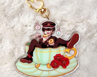 May include: A clear acrylic keychain featuring an anime character in a teacup. The character, wearing a black uniform and hat, is holding a donut. The teacup is light green with red flowers. A gold-colored bunny-shaped clasp is attached.