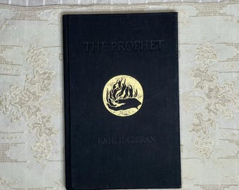 May include: A hardcover book titled "THE PROPHET" by Kahlil Gibran. The book has a black cover with gold lettering and a gold circular emblem. The book is resting on a white and beige patterned surface.