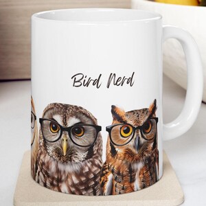 Könnte beinhalten: Weiße Keramiktasse mit einem Design aus drei Eulen mit Brillen. Die Eulen sind braun, schwarz und orange. Die Tasse hat einen schwarzen Henkel und den Text "Bird Nerd" auf der Vorderseite.