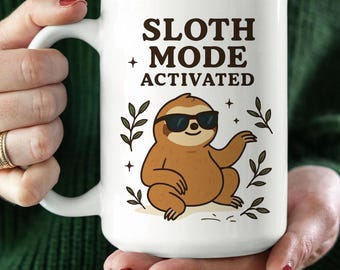 May include: White ceramic mug with the text "Sloth Mode Activated" and a cartoon sloth wearing sunglasses. The sloth is brown with black sunglasses. The mug has a large handle and is held by a person.