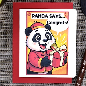 May include: A greeting card with a cartoon panda holding a gift box. The card reads "PANDA SAYS... Congrats!" in a speech bubble. The panda is wearing a red outfit and a hat. The card is on a red envelope.