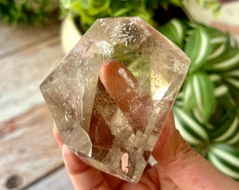 May include: A polished, multifaceted smoky quartz crystal with a translucent, brownish-grey hue. The stone features internal fractures and natural inclusions that catch the light, creating a complex, geometric shape with sharp, defined edges.
