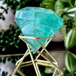 Può includere: Una gemma sfaccettata a forma di diamante color verde acqua, poggiata su un supporto in filo metallico color oro. La pietra presenta una consistenza cristallina traslucida con inclusioni interne, mentre il supporto è costruito con sottili aste metalliche angolari.