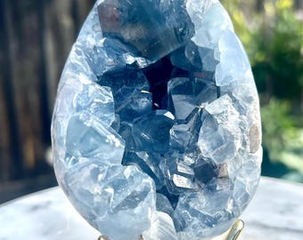 May include: A teardrop-shaped celestite geode featuring a cluster of translucent, pale blue crystalline structures. The mineral specimen is displayed on a simple, gold-toned metal tripod stand, showcasing its natural, jagged interior facets.