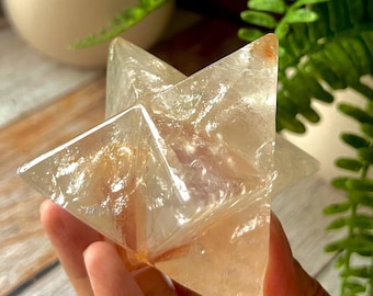 May include: A clear, star-shaped crystal with amber and white hues is held in a hand. The crystal's facets catch the light, creating a sparkling effect. Green plants are visible in the background.