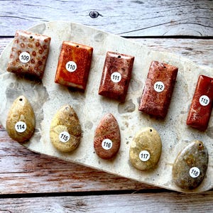 May include: A selection of rectangular and oval stone pendants in brown, red, and beige hues. Each pendant features a unique pattern and is numbered with a white sticker. The pendants are arranged on a speckled, oval-shaped tray.
