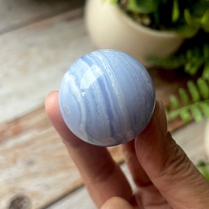 Puede incluir: Una esfera azul claro con bandas blancas y azules, sostenida en una mano. La piedra pulida tiene una superficie lisa y reflectante. El fondo incluye vegetación y una maceta blanca, lo que sugiere un tema natural o decorativo.