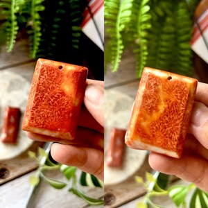 May include: Rectangular coral pendant with a hole for stringing. The pendant has a vibrant orange and white colour with a detailed, textured pattern. The pendant is held in a hand, with a blurred background.