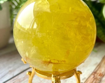 May include: A polished, translucent yellow crystal sphere resting on a decorative gold-toned metal tripod stand. The sphere features internal fractures and inclusions that catch the light, creating a bright, vibrant, and sunny appearance.
