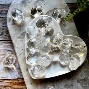 May include: Oval-shaped clear quartz crystal stones are arranged on a white heart-shaped dish and a grey marble-effect surface. The transparent crystals sparkle, reflecting light. The composition highlights natural elements, with a focus on the crystals' clarity.
